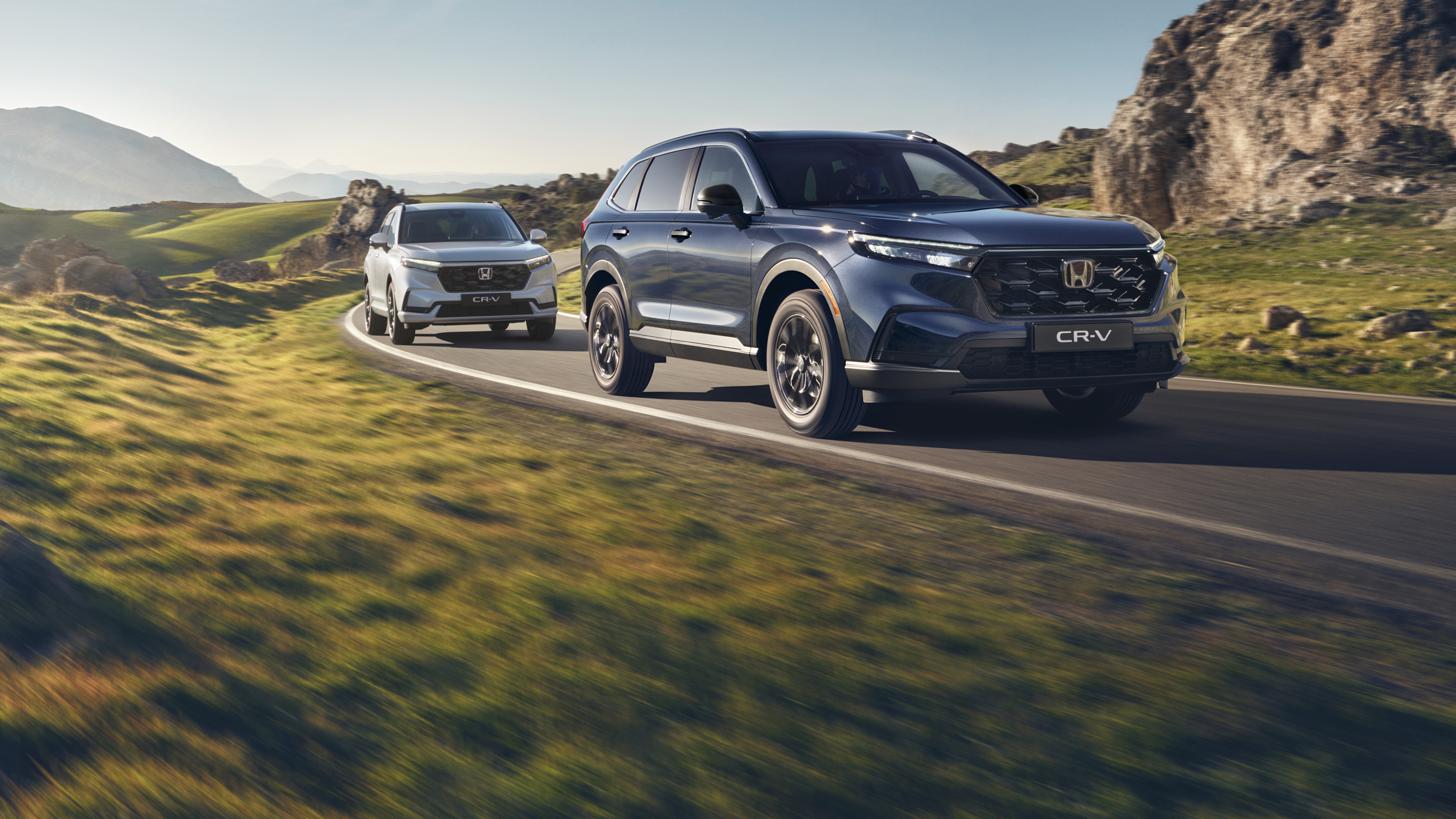 Stříbrné SUV Honda CR-V jedoucí po klikaté horské silnici s kamenitým terénem a horami v pozadí. Vozidlo je v pohybu, což vytváří pocit rychlosti a dobrodružství v drsné krajině.