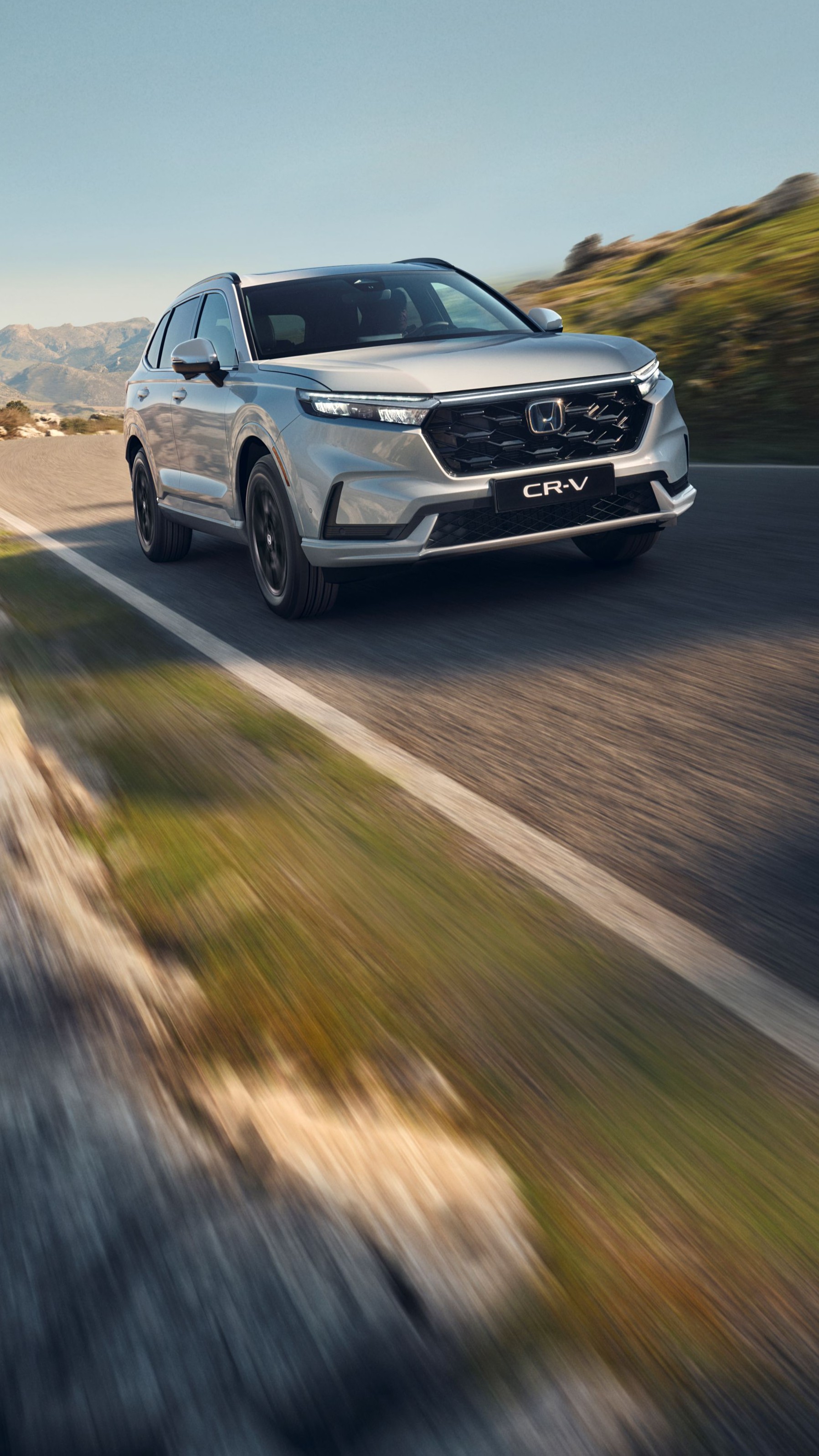 Stříbrné SUV Honda CR-V jedoucí po klikaté horské silnici s kamenitým terénem a horami v pozadí. Vozidlo je v pohybu, což vytváří pocit rychlosti a dobrodružství v drsné krajině.