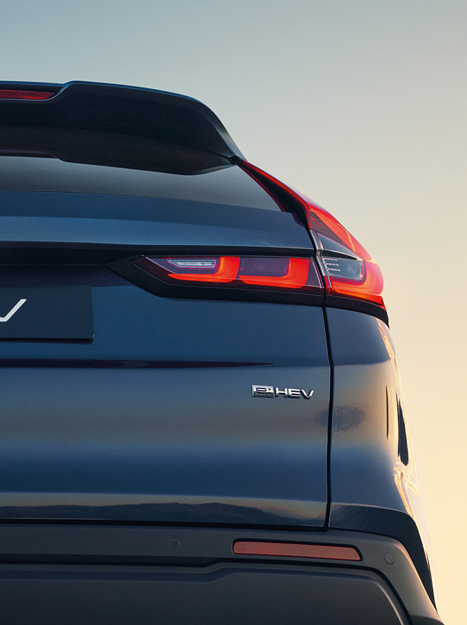 Detailní pohled na zadní část tmavě modrého vozu Honda CR-V e:HEV, na které je vidět znak Honda, nápis CR-V a osvětlená charakteristická zadní LED světla na pozadí jemné oblohy při západu slunce.