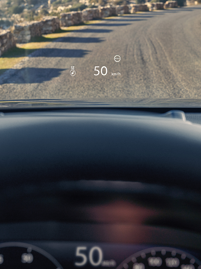 Pohled z místa řidiče, kde je na čelním skle nad štěrkovou cestou promítán Head-Up Display (HUD) s údajem „50 km/h“ a kompasem.
