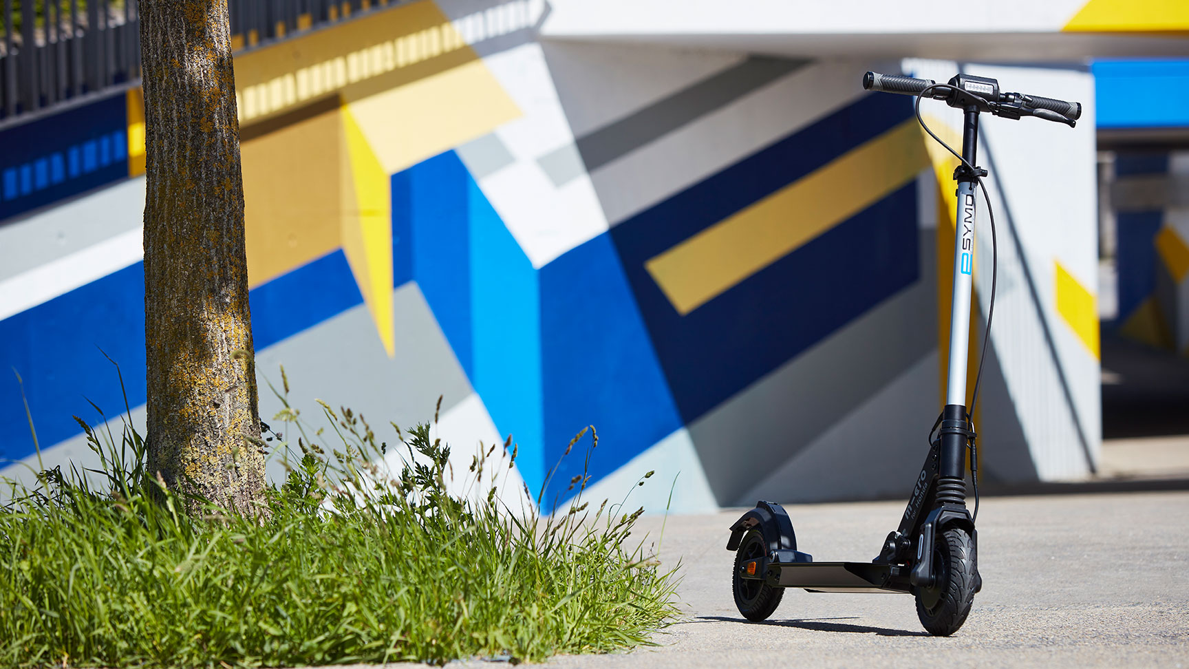 Pohled na koloběžku Honda eSYMO u stromu před pestře pomalovanou zdí