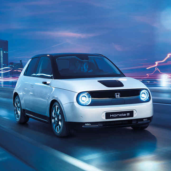 Přední tříčtvrtinový pohled zprava na model Honda e projíždějící elektrickým panoramatem města.