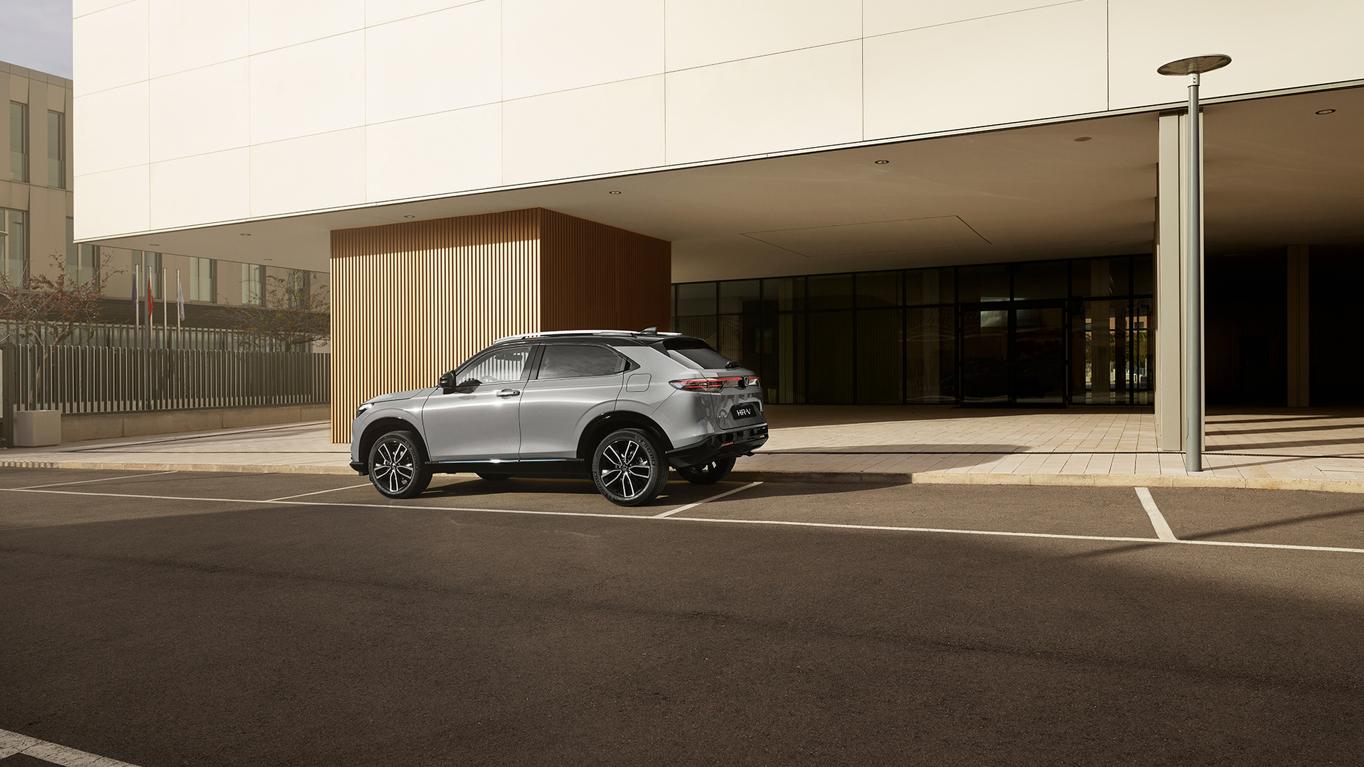 Stříbrná Honda HR-V je zaparkovaná na venkovním parkovišti před moderní budovou s prosklenými stěnami a svislým dřevěným obložením. Vyfocena ze zadního tříčtvrtečního úhlu za denního světla.