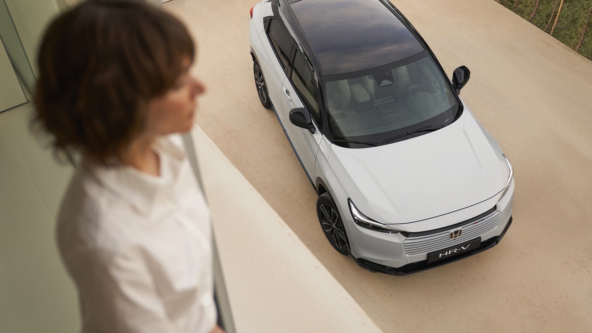Pohled shora na bílý vůz Honda HR-V s černou střechou zaparkovaný na světlé příjezdové cestě. V popředí uvnitř stojí rozostřená postava a dívá se dolů směrem k vozu.