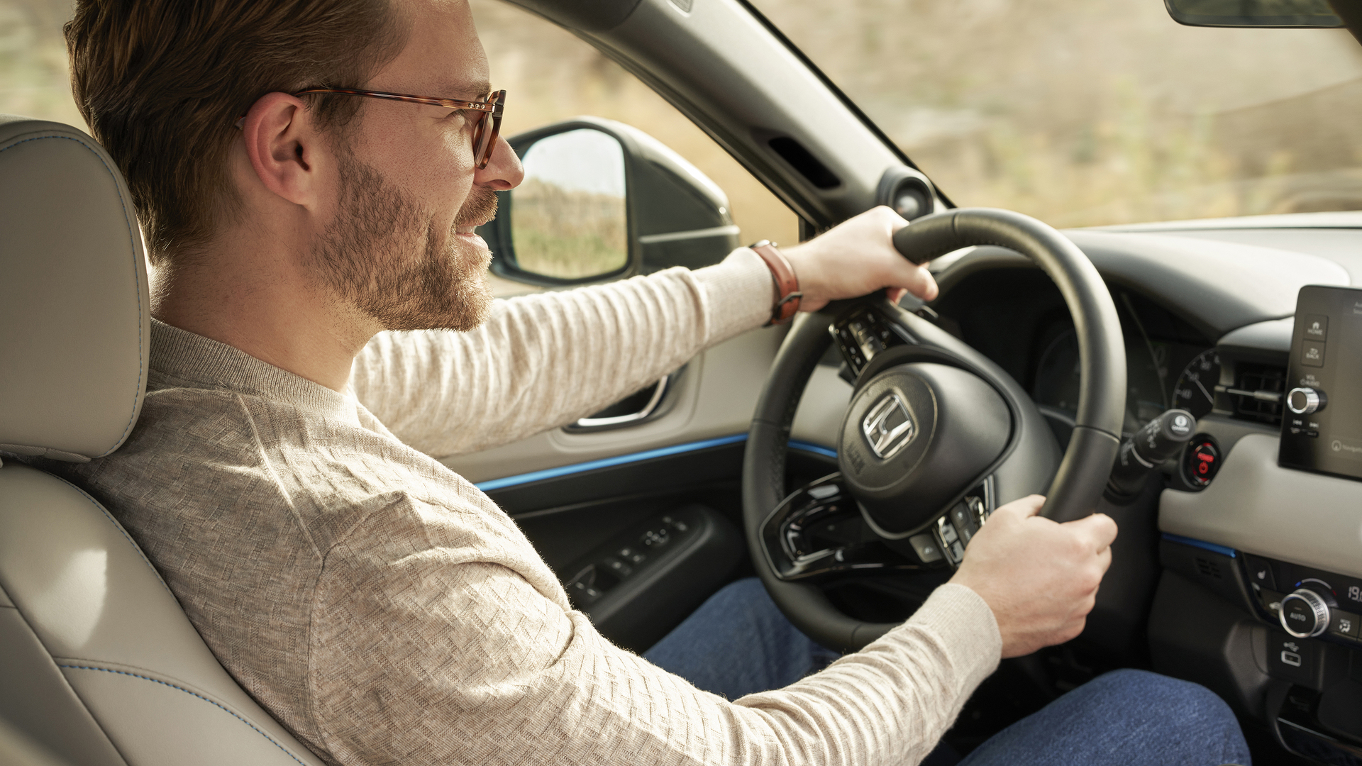 Muž s brýlemi řídí vůz Honda HR-V; pohled ze sedadla spolujezdce; obě ruce má na volantu a palubní deska je viditelná.