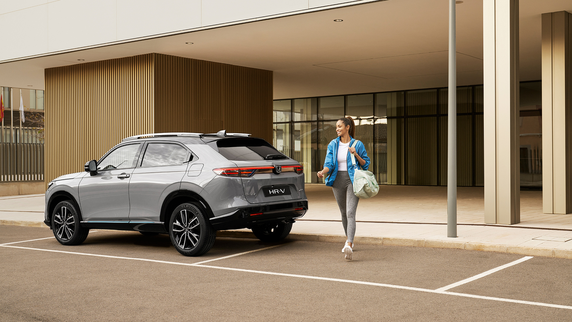 Stříbrná Honda HR-V zaparkovaná před moderní budovou, zatímco kolem ní prochází žena s taškou, což naznačuje každodenní městské využití a praktičnost.