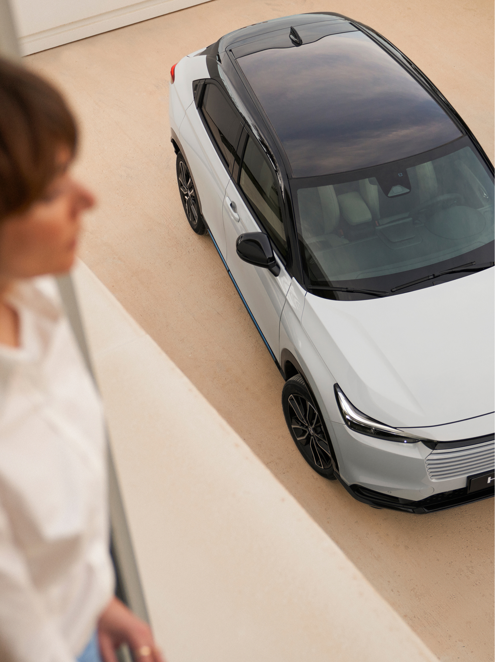 Pohled shora na bílý vůz Honda HR-V s černou střechou zaparkovaný na světlé příjezdové cestě. V popředí uvnitř stojí rozostřená postava a dívá se dolů směrem k vozu.