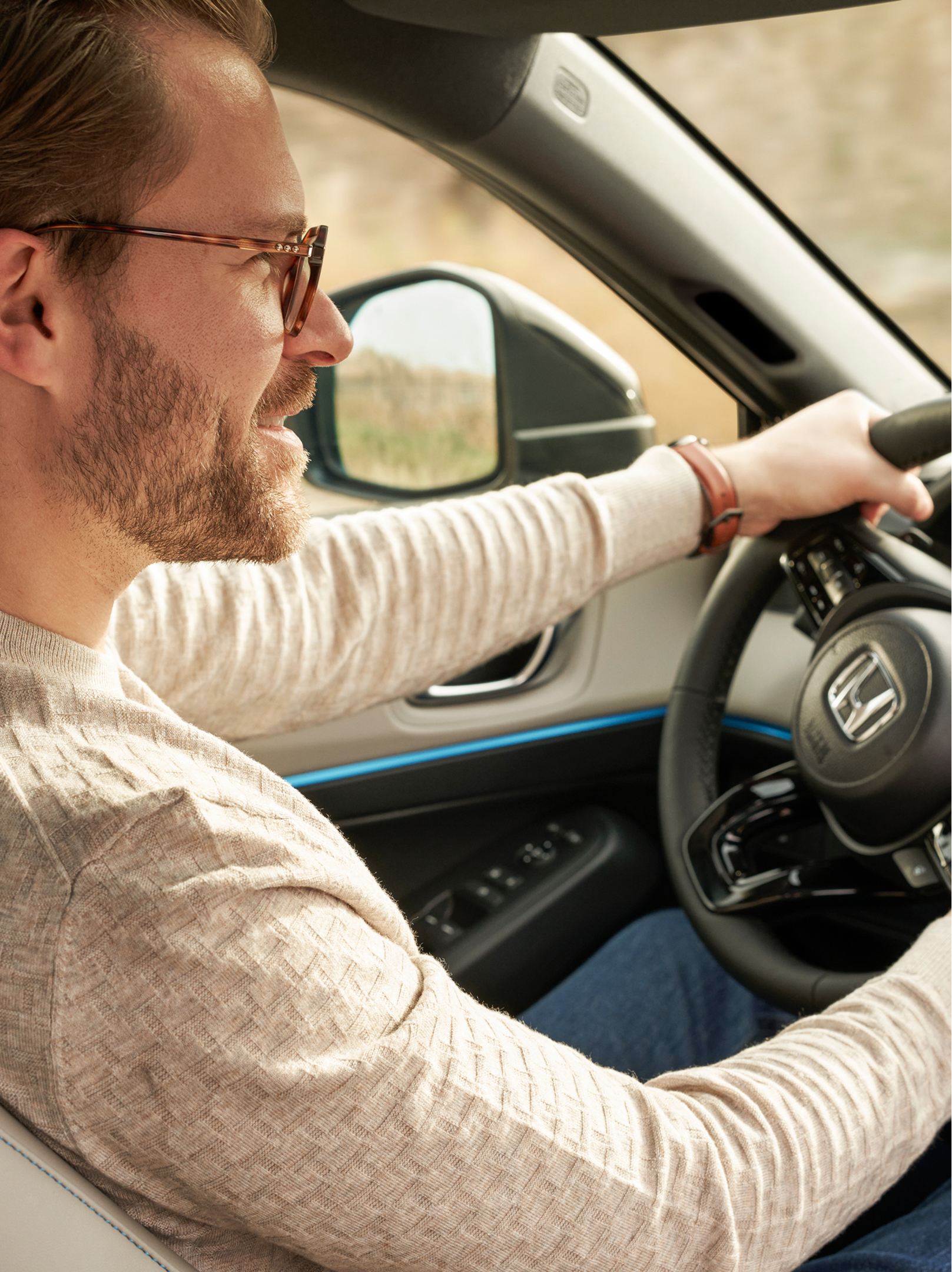 Muž s brýlemi řídí vůz Honda HR-V; pohled ze sedadla spolujezdce; obě ruce má na volantu a palubní deska je viditelná.
