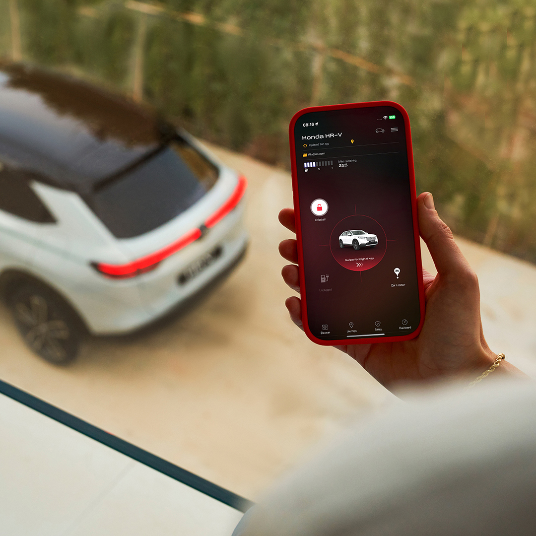 Osoba drží chytrý telefon zobrazující aplikaci pro nastavení nabíjení vozidla, zatímco v pozadí je zaparkované bílé SUV.