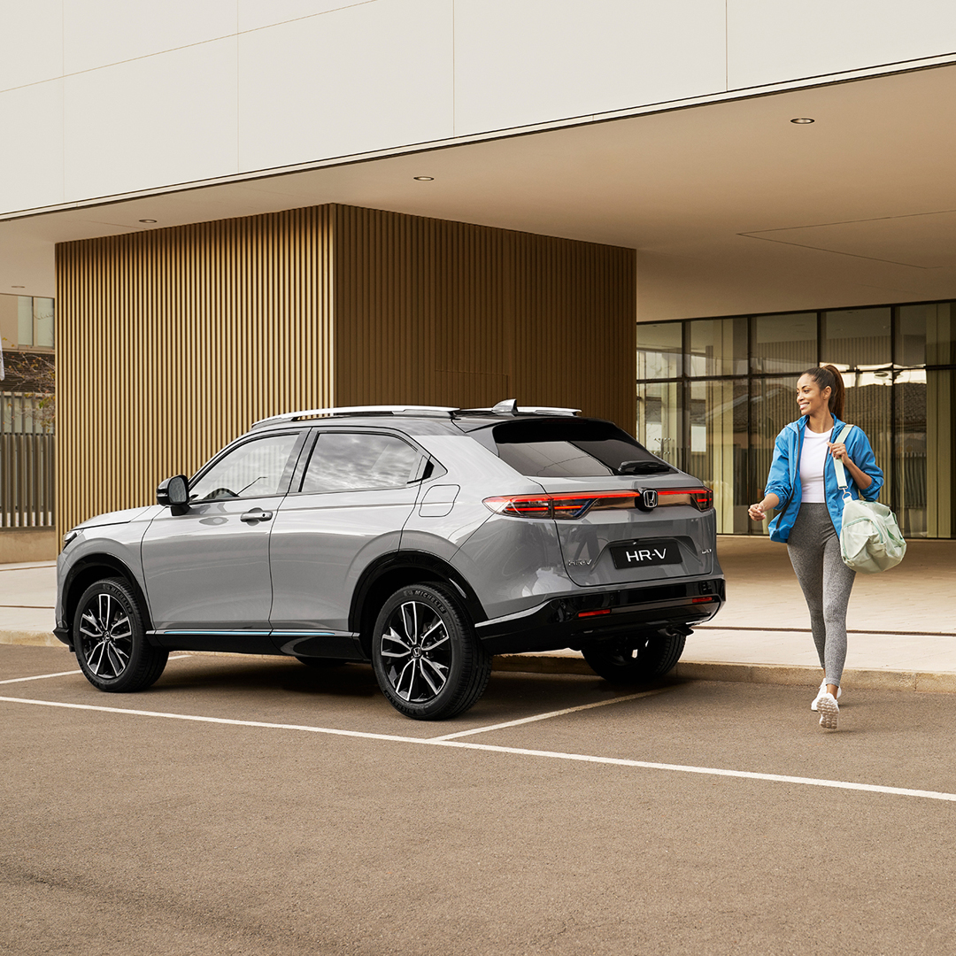 Stříbrná Honda HR-V zaparkovaná před moderní budovou, zatímco kolem ní prochází žena s taškou, což naznačuje každodenní městské využití a praktičnost.