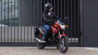Model Honda CB125F v červeném provedení, přední tři čtvrtiny, pohled z pravé strany, jezdec na stojícím motocyklu, ve městě