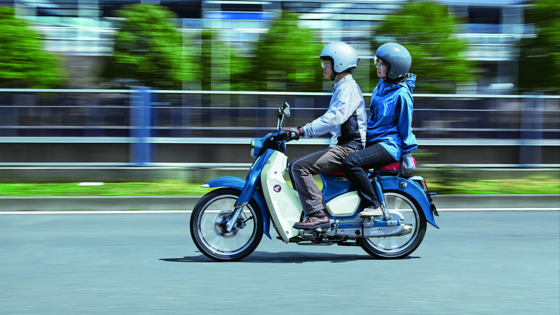 A Honda Super Cub motorcycle in motion on a city street, carrying two riders wearing helmets. The scooter is blue and white, and the background is blurred, suggesting movement.