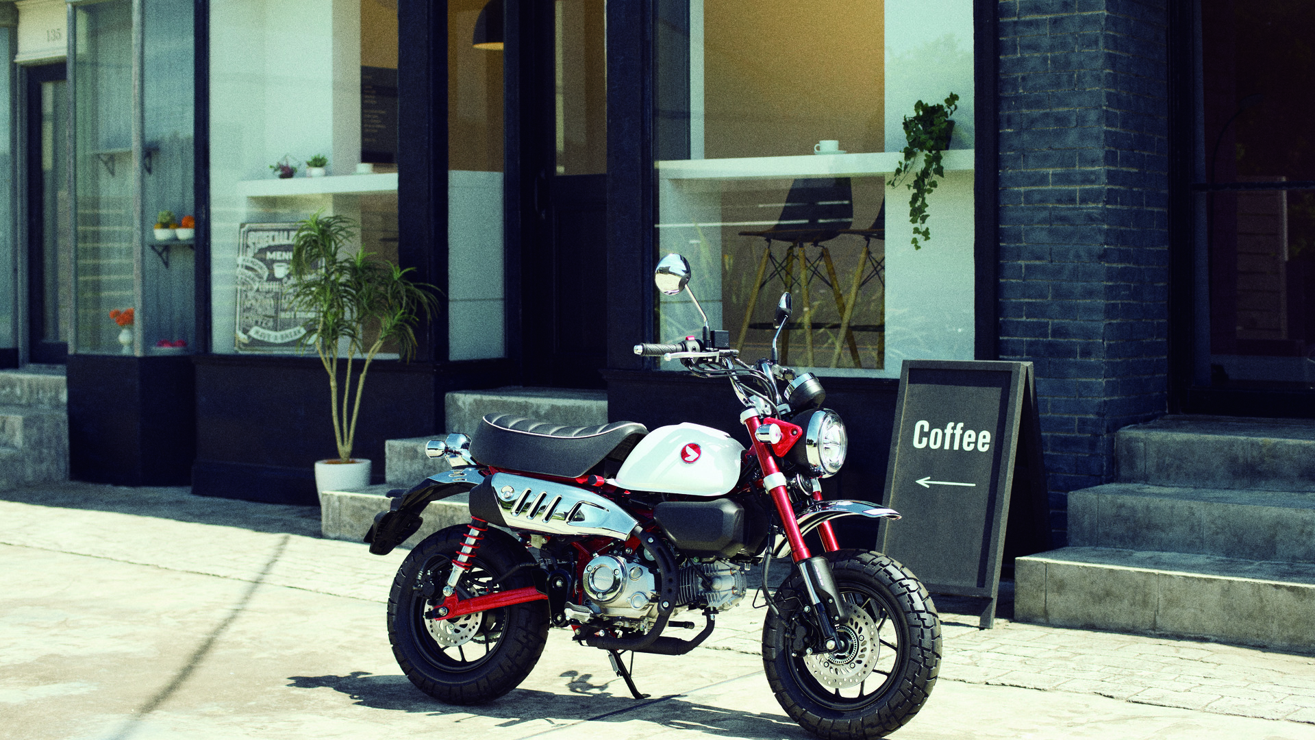 A white Honda Monkey mini motorcycle parked on a sidewalk in front of a cafe or coffee shop. The small, retro-styled bike is positioned near a chalkboard sign advertising coffee.