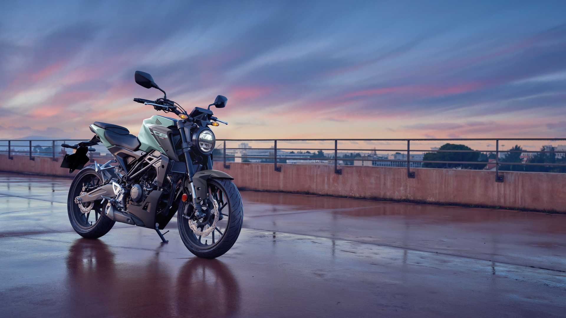 A Honda CB300R motorcycle parked on a wet rooftop or elevated platform against a colorful sunset sky. The sleek, modern bike is positioned facing slightly to the right, with its metallic body reflecting the ambient light. In the background, a railing and cityscape are visible, creating a dramatic urban setting.