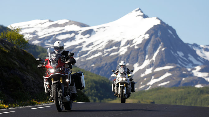 Dva motocykly jedoucí po silnici se zasněženými horami v dálce.