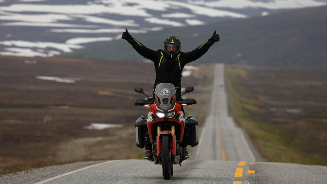 Muž na motocyklu Honda Africa Twin se zvednutýma rukama na silnicích v Nordkappu.