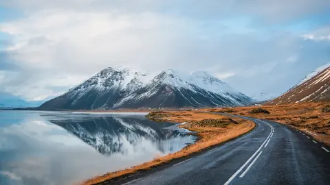 Zimní panoramatická fotografie silnice vedoucí podél pobřeží jezera k vulkanickým horám. Vysoké skalnaté vrcholy pokryté vrstvou sněhu zrcadlící se na vodní hladině. Pohled řidiče na Islandský silniční okruh.