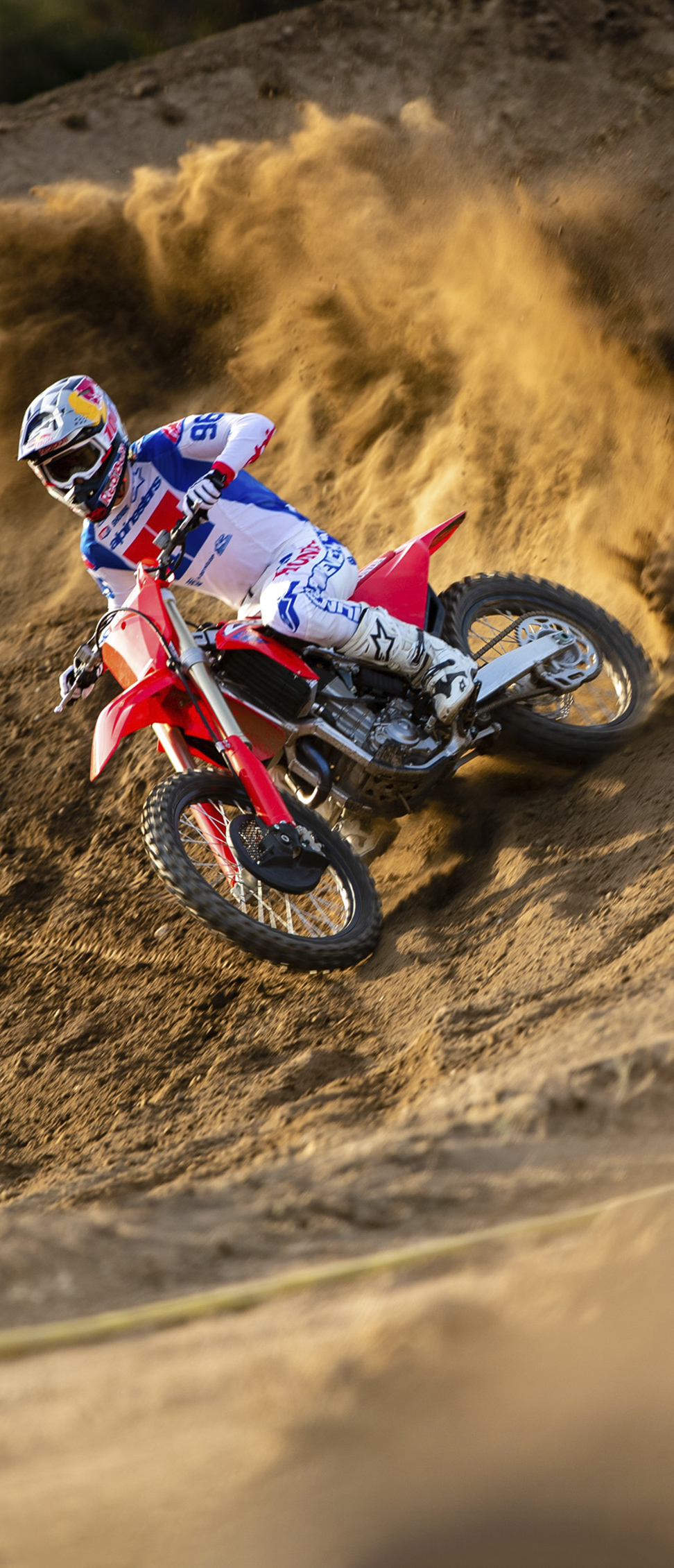 Motokrosový jezdec v bílé, modré a červené výstroji naklání červený terénní motocykl do strmého písečného násypu a při průjezdu zatáčkou vymrětí do vzduchu velkou vlnu písku.