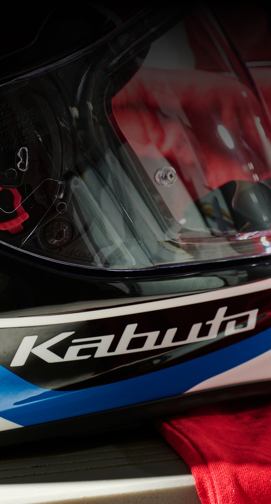Close-up view of a motorcycle helmet visor with the \"Kabuto\" brand name visible on the lower edge. The helmet appears to be black with blue and white accents.