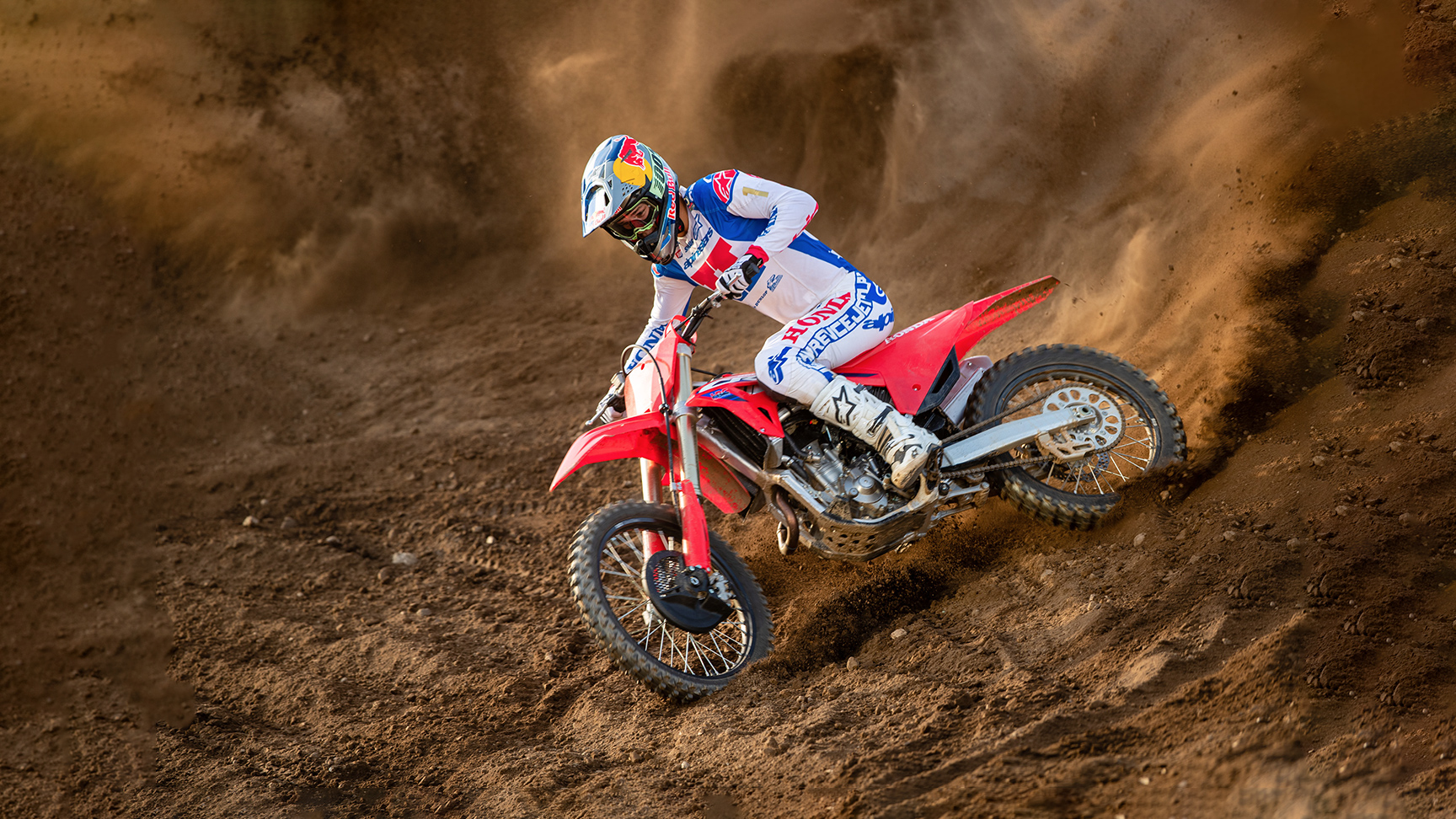 Motokrosový jezdec v bílé, modré a červené závodní výstroji se na červeném terénním motocyklu prudce naklání do zatáčky, projíždí písečnou tratí a víří za sebou dramatický oblak písku.