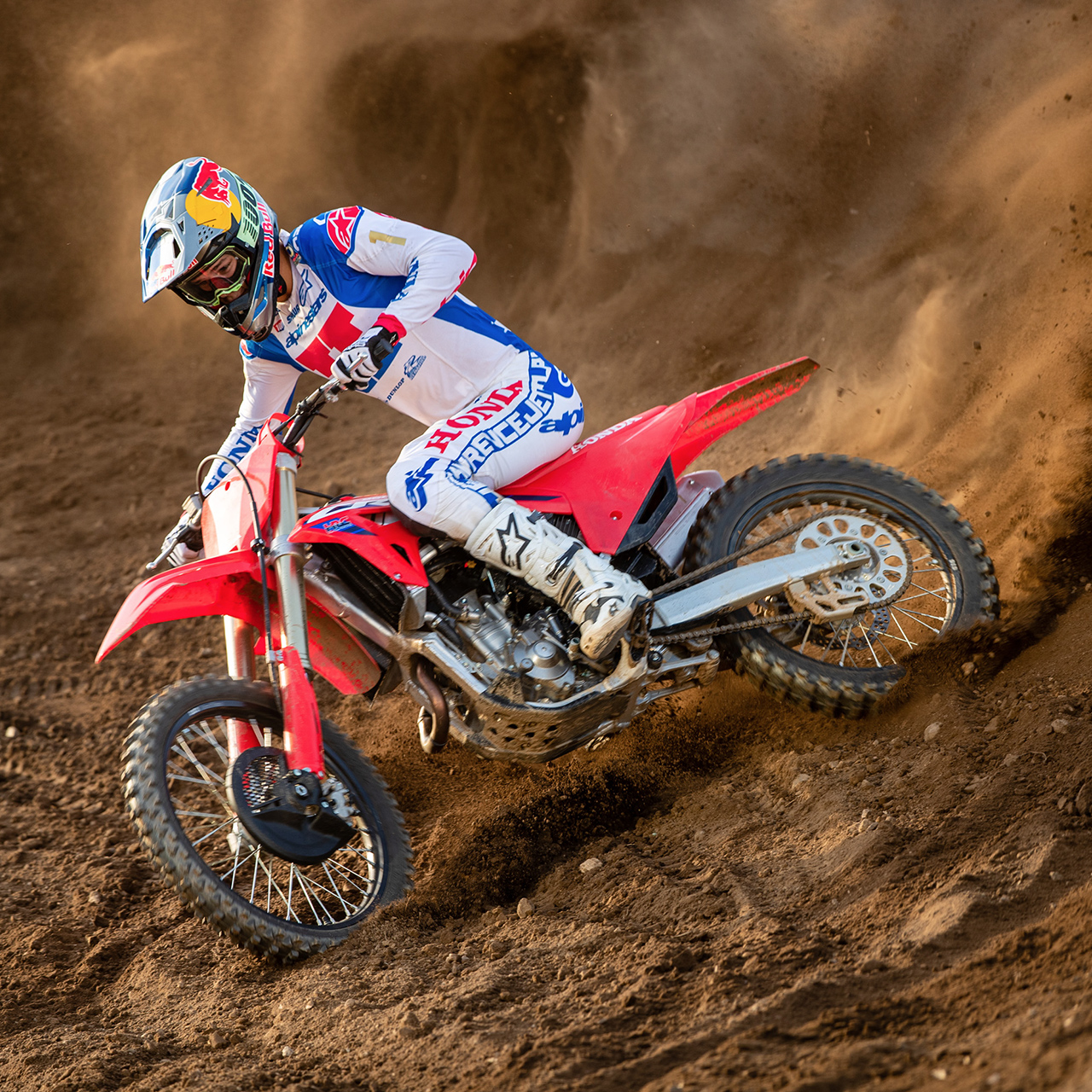 Motokrosový jezdec v bílé, modré a červené závodní výstroji se na červeném terénním motocyklu prudce naklání do zatáčky, projíždí písečnou tratí a víří za sebou dramatický oblak písku.