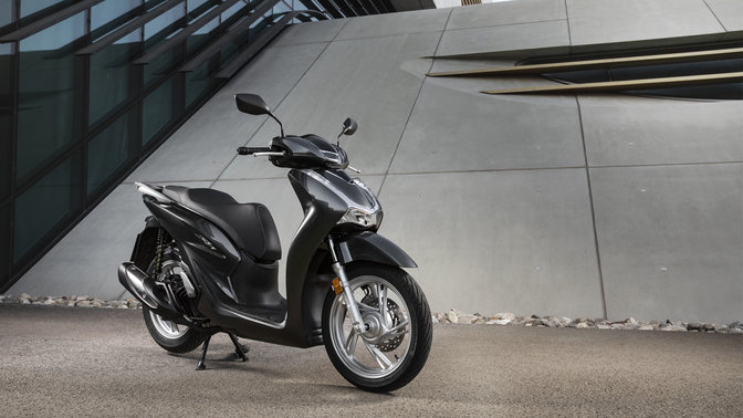 Honda Sh125i s horním boxem a čelním štítem na ulici.