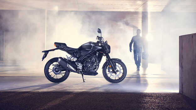 Boční pohled na motocykl Honda CB300R zaparkovaný v garáži s mužem jdoucím k motocyklu.